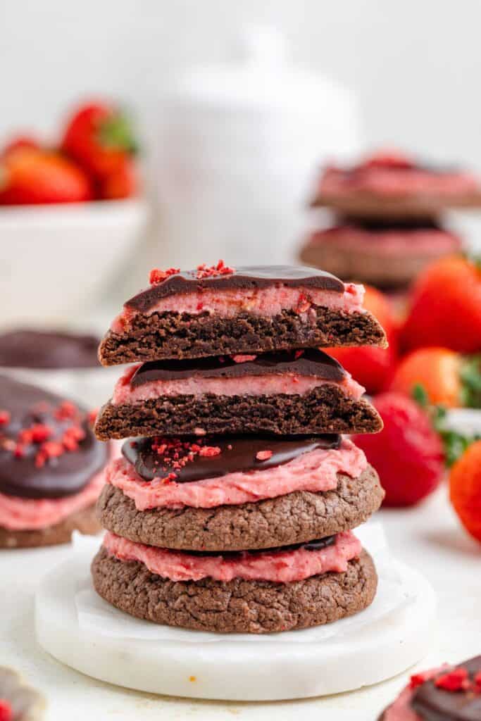 Stack of chocolate and strawberry cookies with pink frosting and chocolate topping, surrounded by strawberries and extra cookies.