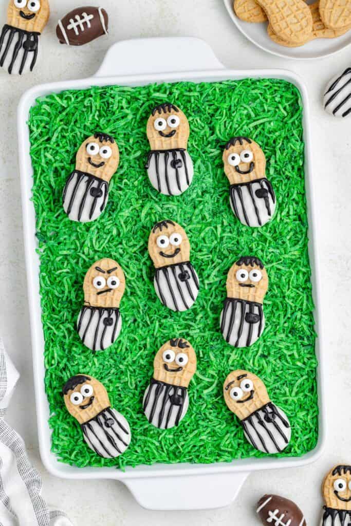 Cookies decorated as football referees on green coconut "field," with mini football-shaped cookies nearby.