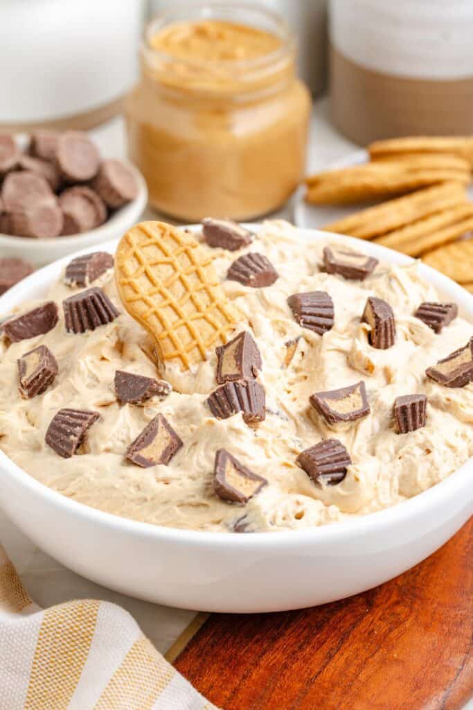 Creamy peanut butter dessert dip topped with chopped peanut butter cups, served with cookies; jars and candies in the background.