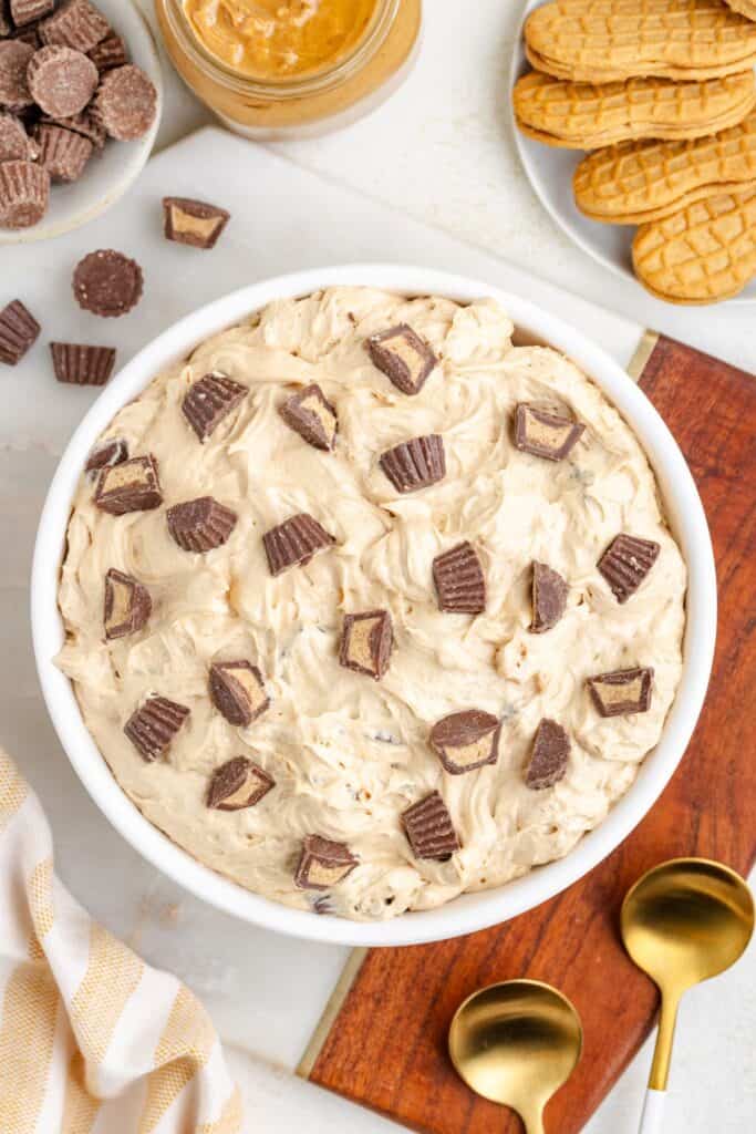 Bowl of creamy peanut butter dip with mini chocolate peanut butter cups, surrounded by cookies and a peanut butter jar.