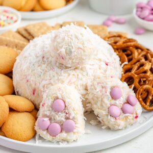 Bunny tail-shaped coconut dessert with pink candy paws, surrounded by cookies and pretzels on a white plate.