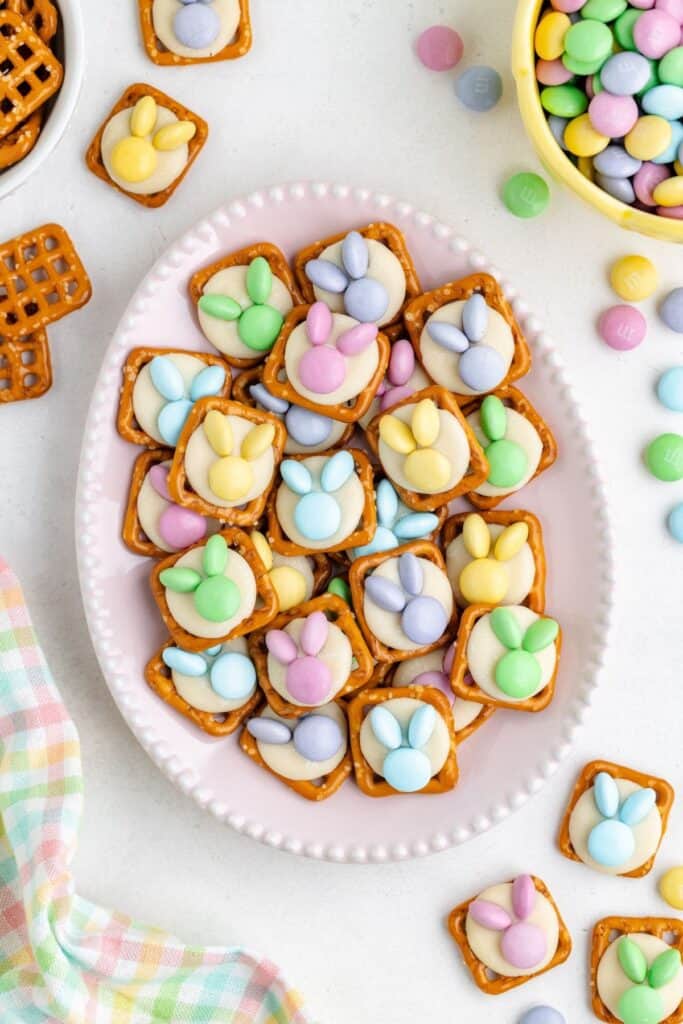 Plate of Easter pretzels decorated as bunny faces with white chocolate and candy, surrounded by colorful candies.