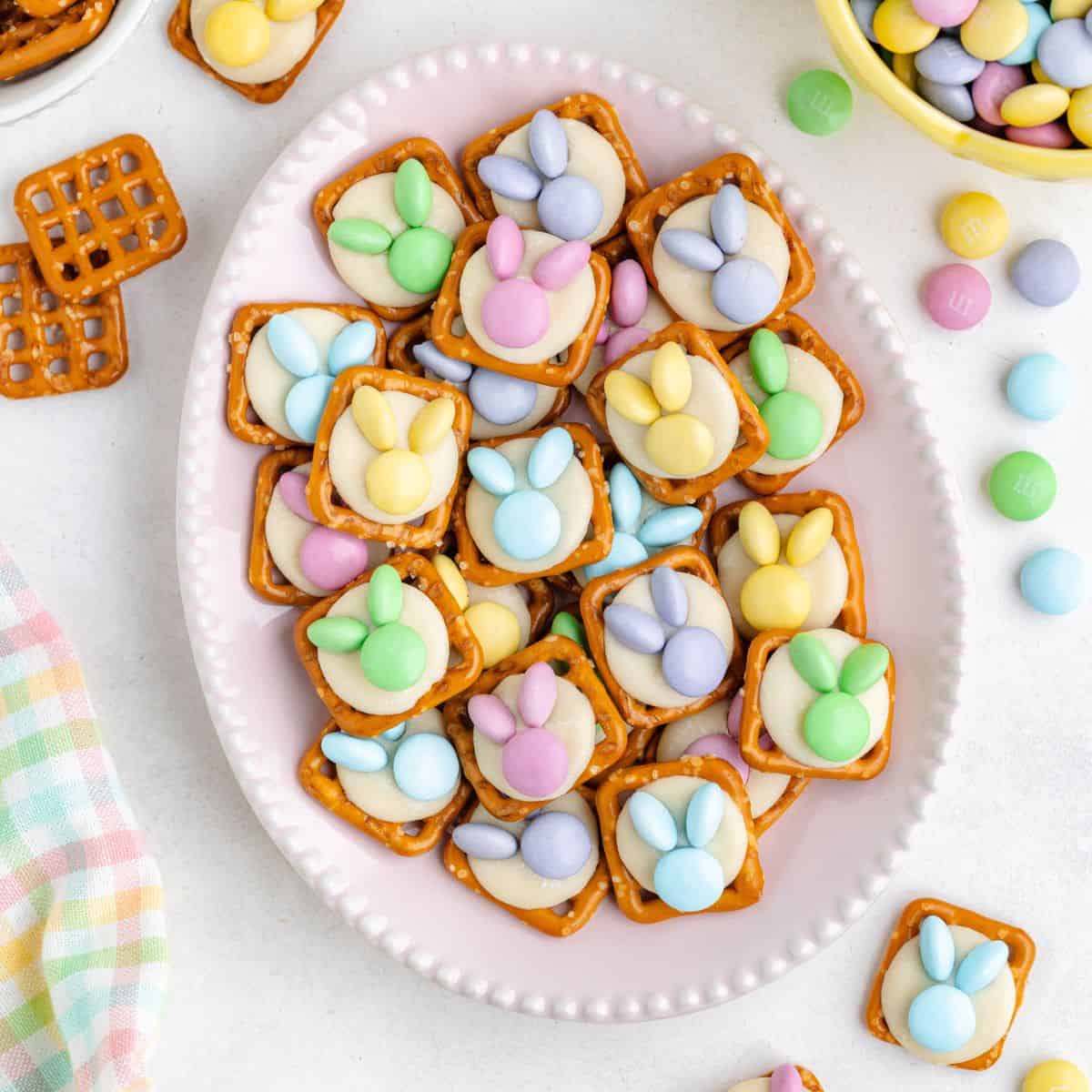 Pink plate with bunny pretzel topped with white chocolate and Easter M&Ms; candies and pretzels nearby.