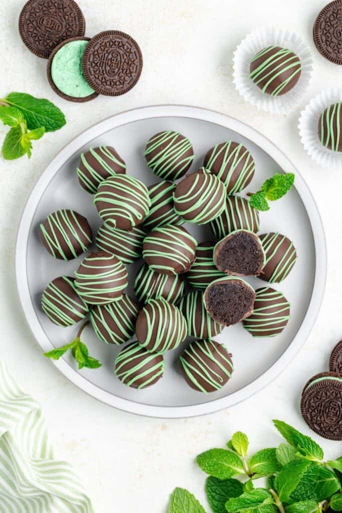 Plate of chocolate mint truffles with green drizzle, surrounded by mint leaves and cookies on a white surface.