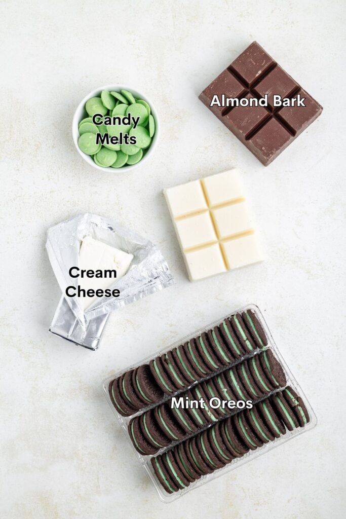 Five dessert ingredients—green candy melts, chocolate and white almond bark, cream cheese, mint Oreos—on a light surface.