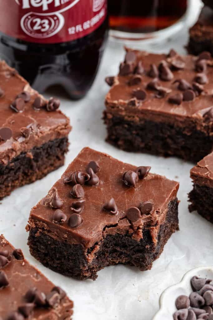 A bitten Dr Pepper brownie with frosting sits among others; Dr. Pepper bottle and glass in background.