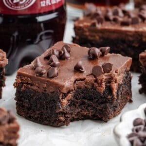 Close-up of a bitten Dr Pepper brownie with frosting and chips; more brownies and a bottle blurred in the background.