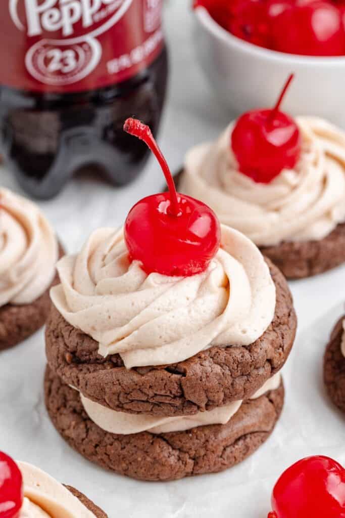 Stack of Dr Pepper cookies with light brown frosting and cherries on top; Dr Pepper bottle and cherries behind.