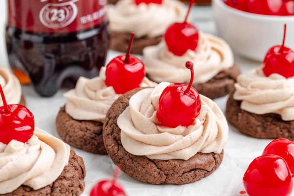 Chocolate Dr Pepper cookies with frosting and cherries on top, soda bottle and bowl of cherries in the background.