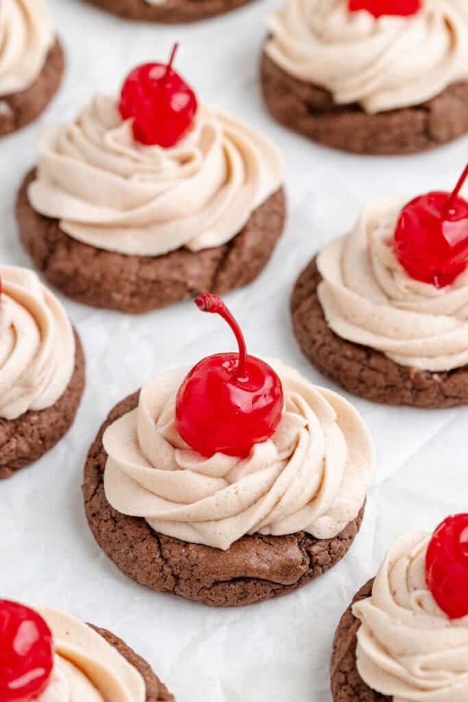 Cake mix Dr Pepper cookies with light brown frosting swirls and maraschino cherries, arranged on a white background.
