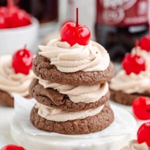 Three stacked Dr Pepper cookies with soda frosting and a cherry on top; more cookies and cherries behind.