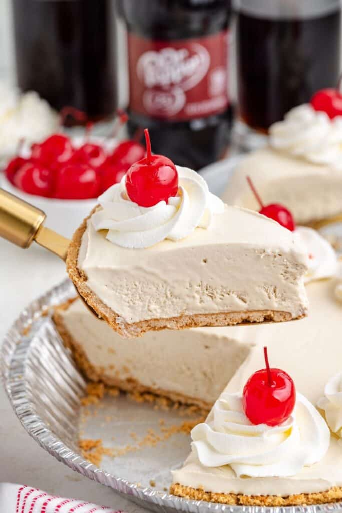 A slice of no bake Dr Pepper pie with whipped cream and a cherry is lifted from the tin; bottles in background.