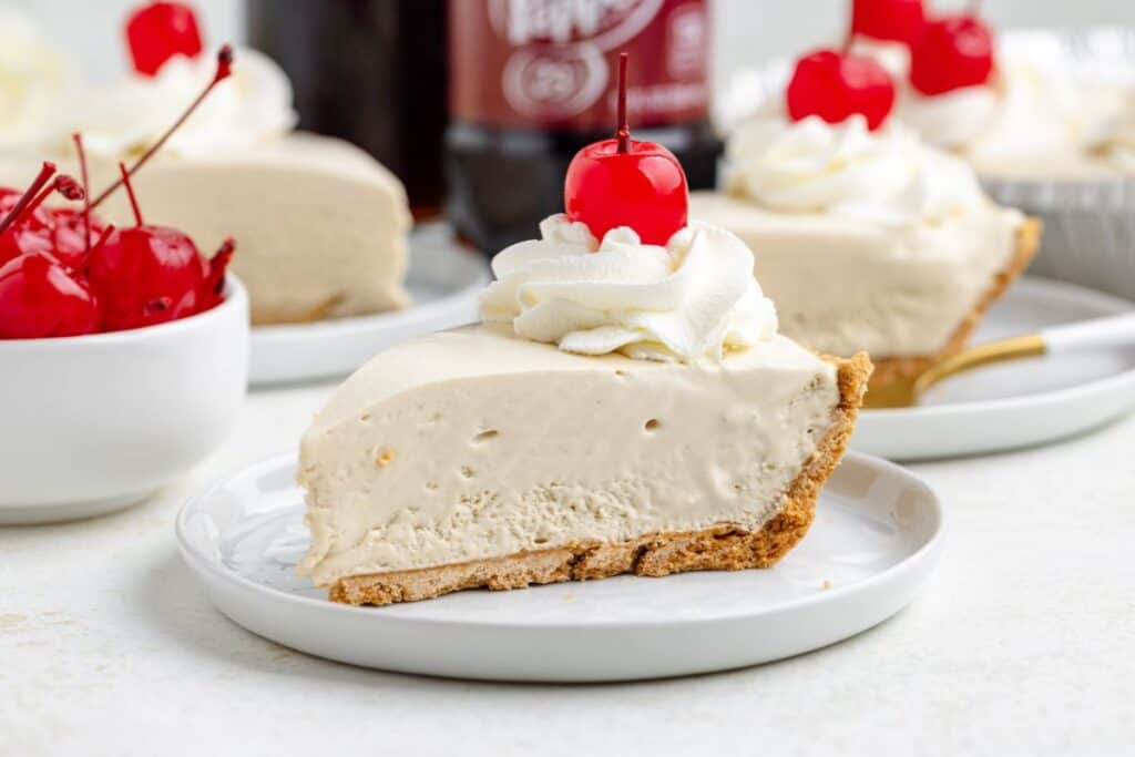 A slice of Dr Pepper float pie with whipped cream and a cherry on a plate; cherries and another slice behind.