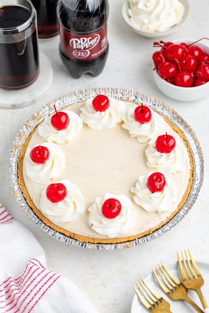 Dr Pepper Pudding Pie topped with whipped cream swirls and cherries sits on a table with various dessert accompaniments.