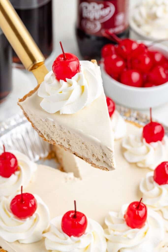 A slice of Dr Pepper pie with whipped cream and a cherry is lifted, with more pie, cherries, and soda bottles behind.