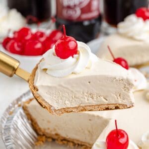 Slice of frozen Dr Pepper pie with graham cracker crust, whipped cream, and cherry being served from a tin.