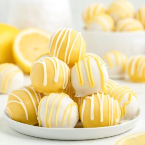 Plate of yellow and white lemon truffles with icing drizzle, sliced lemons and extra truffles in background.