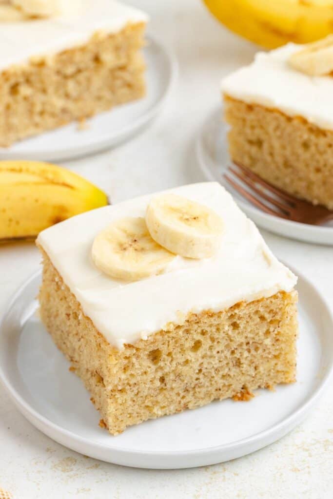 Square slice of cake mix banana cake with cream cheese frosting and banana slices, served on a white plate.