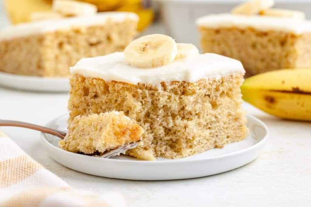Banana cake made with cake mix with cream cheese frosting and banana topping on a plate, fork holds a small bite.
