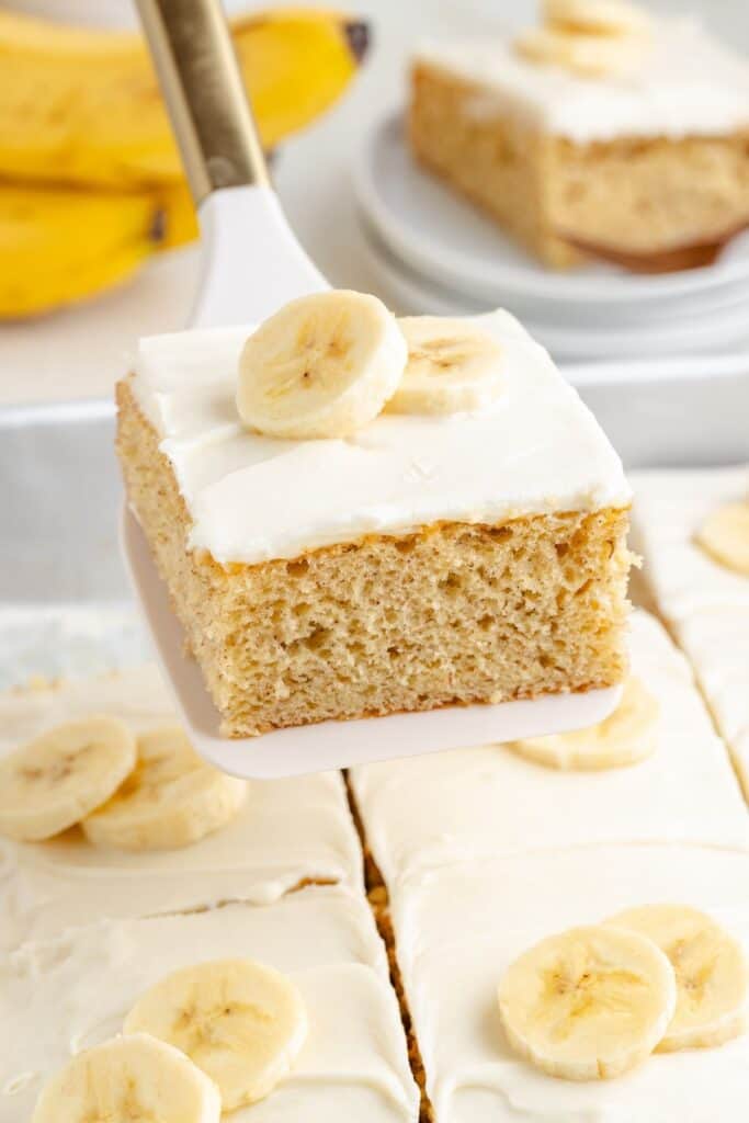 A slice of easy banana cake with cream cheese frosting and banana slices is lifted from a tray; more cake in background.