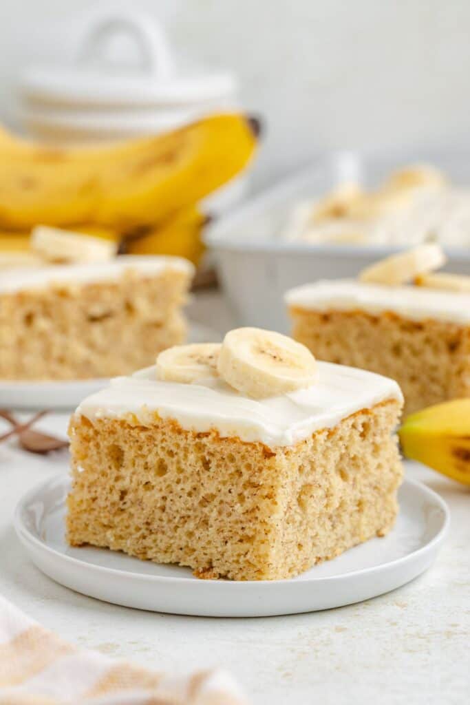 Square banana cake using a cake mix with white frosting and banana slices on a white plate; more cake pieces and a banana behind.