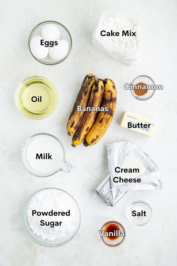 Baking ingredients like eggs, cake mix, bananas, and more arranged neatly on a light-colored surface.
