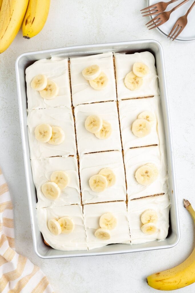 A frosted banana cake cut into twelve squares, each topped with banana slices, in a rectangular pan.