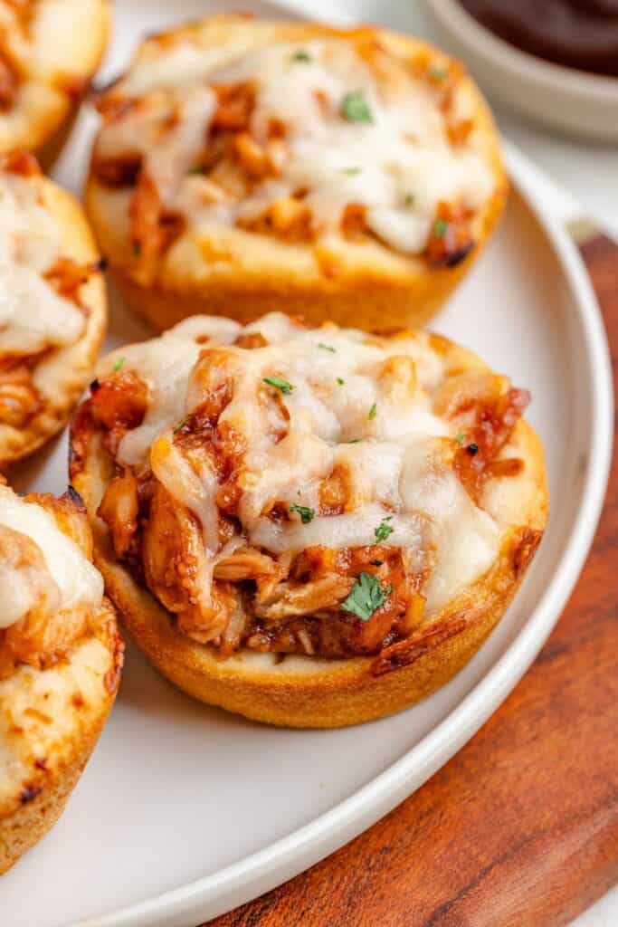 Baked biscuit cups with barbecue chicken and melted cheese, topped with herbs on a white plate.