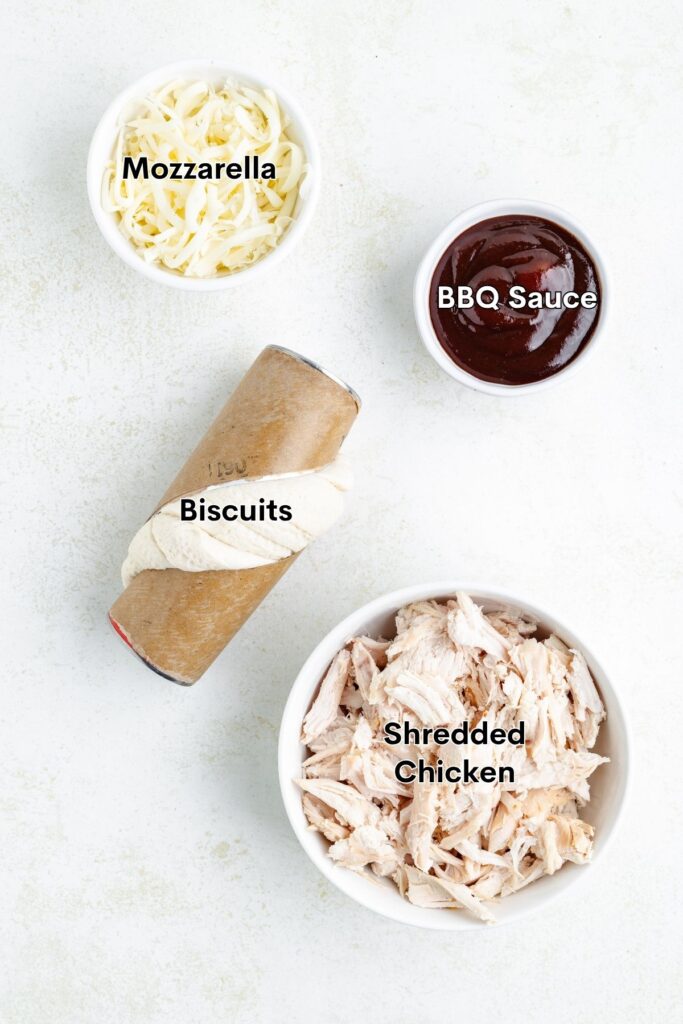 Four labeled ingredients on a white surface: shredded chicken, biscuits, mozzarella cheese, and BBQ sauce.