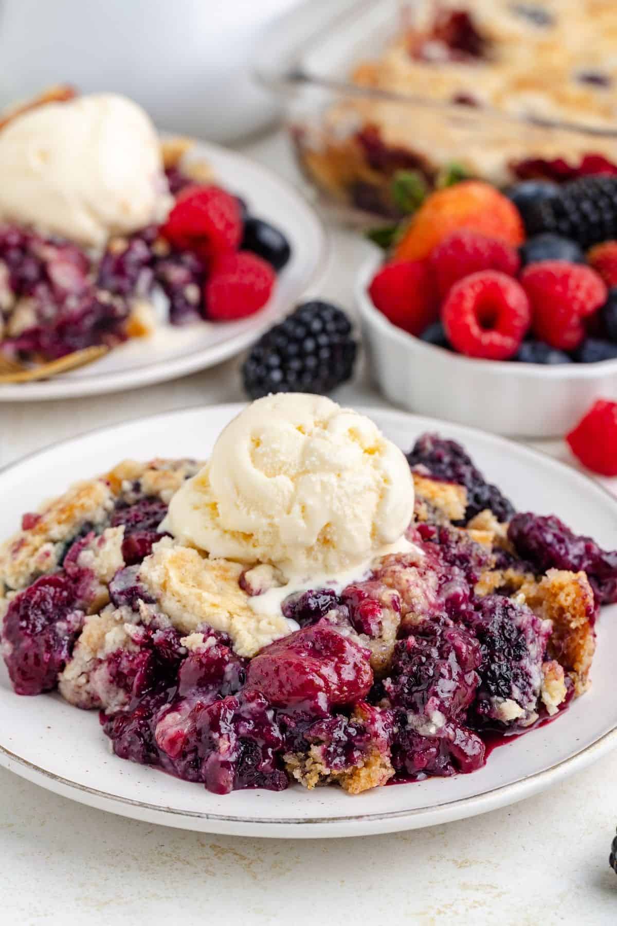 Mixed berry cobbler topped with vanilla ice cream on a plate; fresh berries and more cobbler in the background.