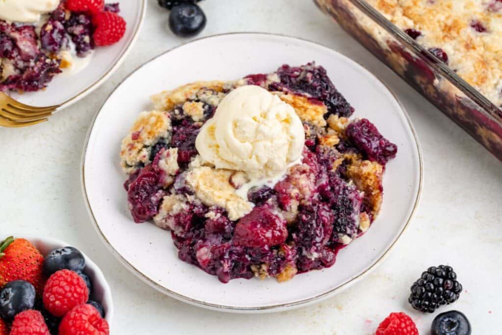 Plate of mixed berry cobbler with vanilla ice cream, fresh berries and baking dish visible in the background.