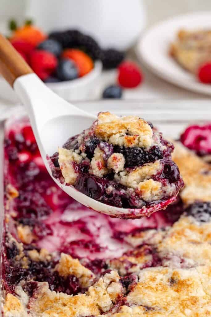 Close-up of mixed berry cobbler being served with a white spoon from a baking dish; berries and cobbler behind.