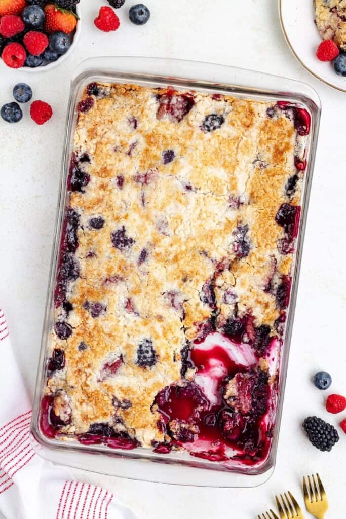 Rectangular glass dish with mixed berry cobbler, a serving scooped out revealing fruit filling and golden crust.