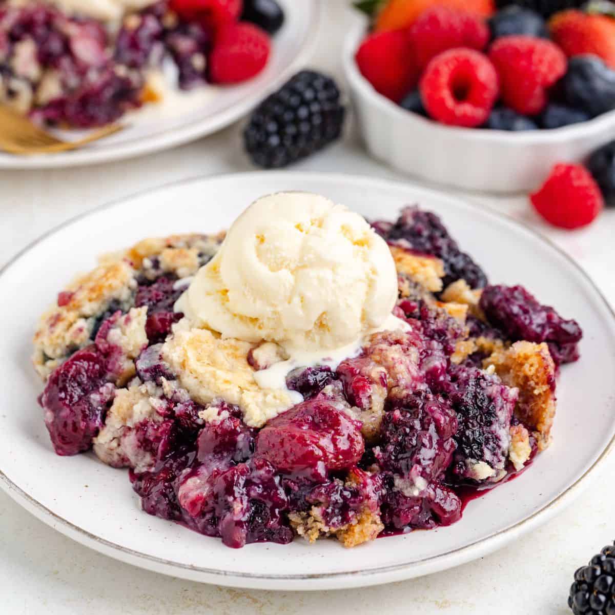 Mixed berry dump cake topped with vanilla ice cream on a plate; fresh berries sit in the background.