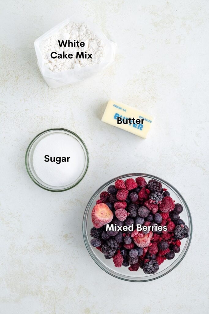Top view of mixed berries, sugar, butter, and cake mix on a light surface, each ingredient clearly labeled.