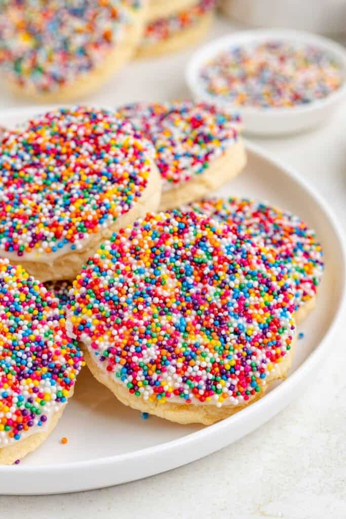 Frosted sprinkle cookies sit on a white plate; a bowl of extra sprinkles is in the background.