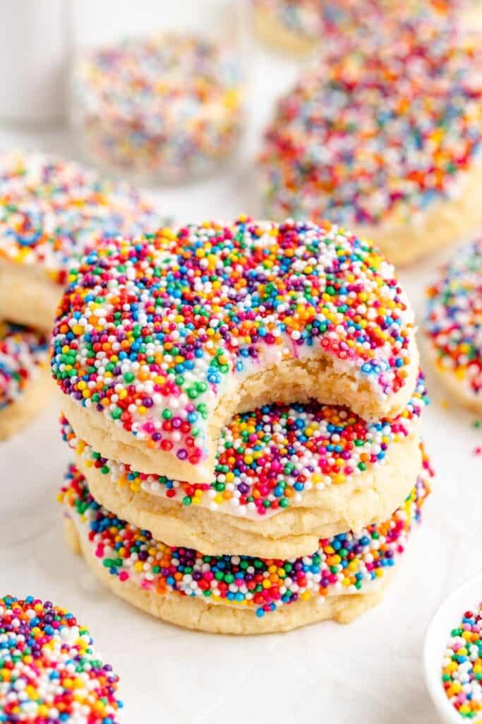 Three frosted cookies with sprinkles; top cookie has a bite. More cookies in background.