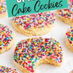 Colorful sprinkle cake mix cookies on parchment; one has a bite missing. Sign reads "Sprinkle Cake Cookies.