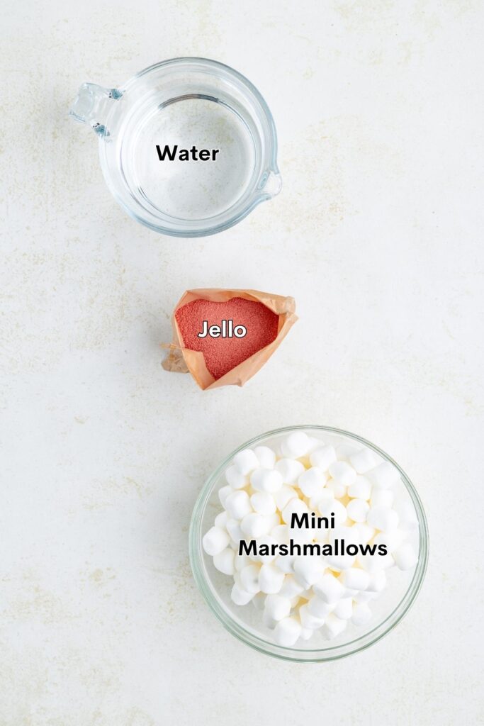 Measuring cup of water, open red Jello packet, and bowl of mini marshmallows on a light surface.