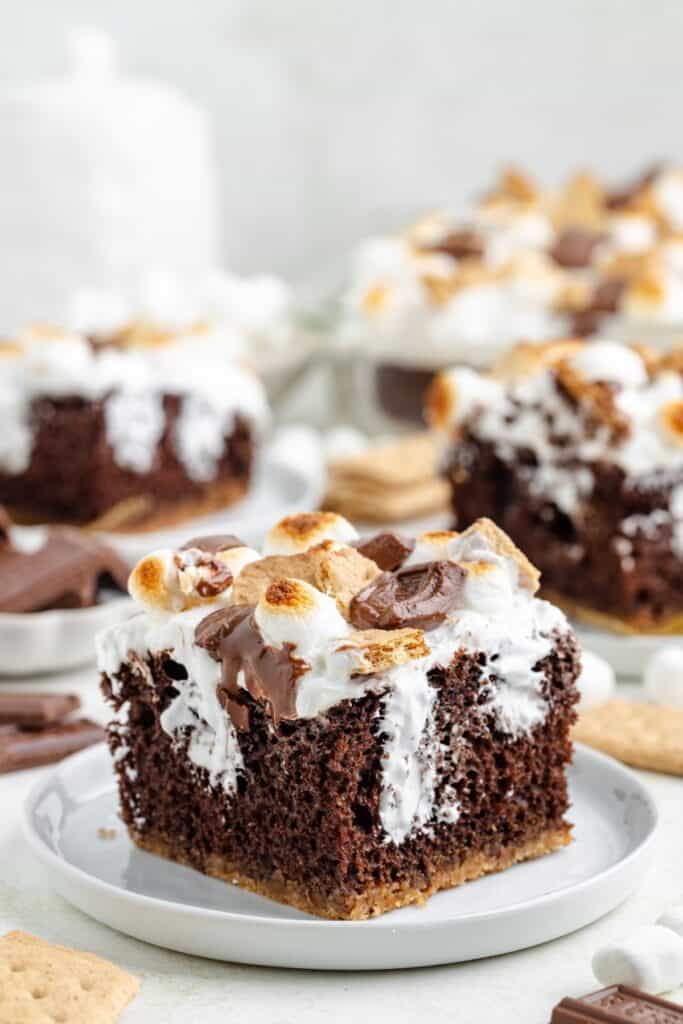 A slice of s'mores poke cake with toasted marshmallows and chocolate on a white plate; more cake in the background.
