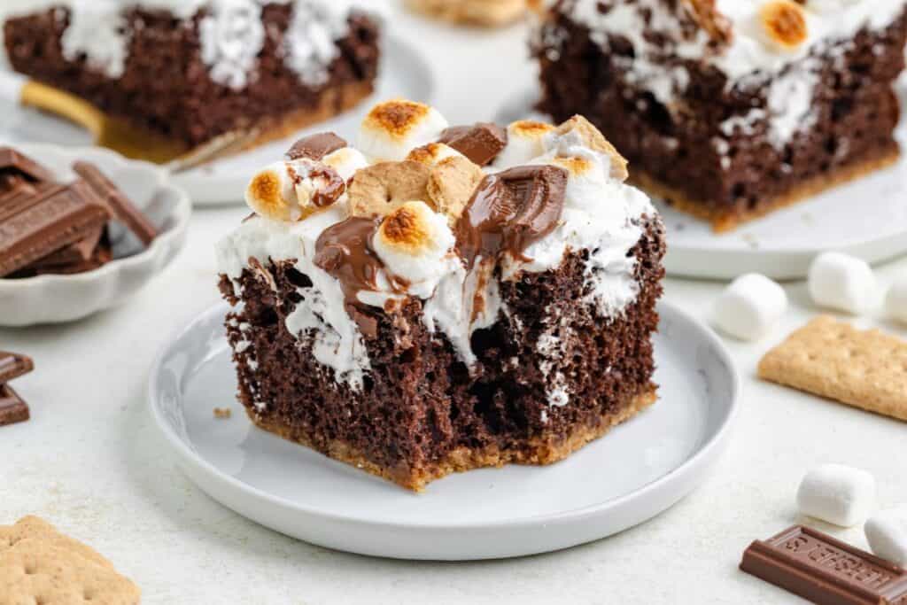 Smores poke cake with toasted marshmallows, melted chocolate, and graham crackers on a white plate.