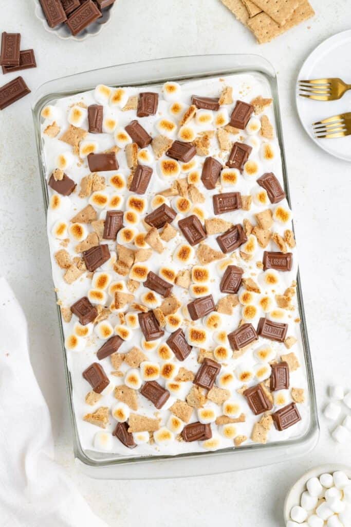 A glass dish of s'mores cake with toasted mini marshmallows, chocolate pieces, and broken graham crackers.