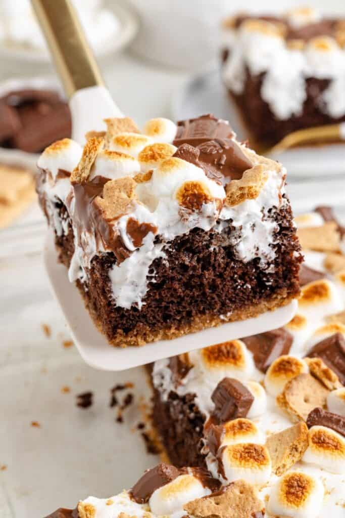 Spatula lifting a cake mix smores cake square topped with toasted marshmallows, chocolate, and graham crackers from a tray.