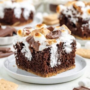 S'mores poke cake with toasted marshmallows and chocolate pieces on a white plate.