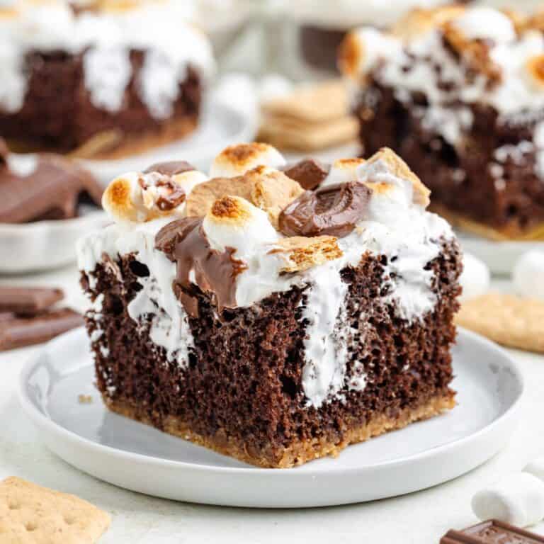 S'mores poke cake with toasted marshmallows and chocolate pieces on a white plate.