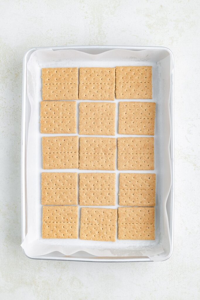 Baking pan with parchment paper holds fifteen rectangular graham crackers, neatly arranged in three rows.