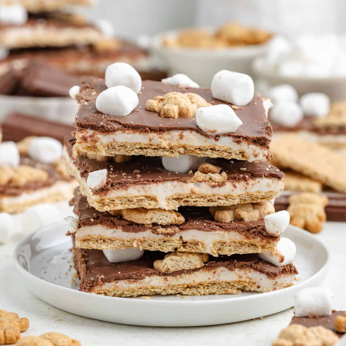 Stacked chocolate s'mores bark on a white plate, surrounded by scattered marshmallows and graham crackers.