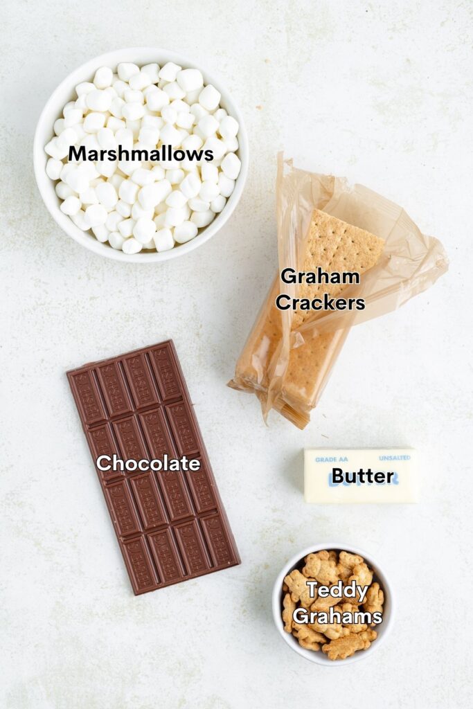 Five labeled ingredients—marshmallows, graham crackers, chocolate, butter, and Teddy Grahams—arranged on a light surface.