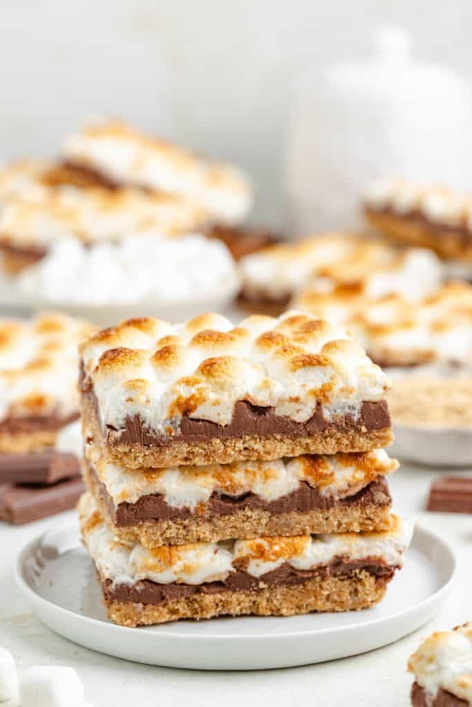 Three stacked s'mores bars on a white plate, with visible layers of crust, chocolate, and toasted marshmallow.
