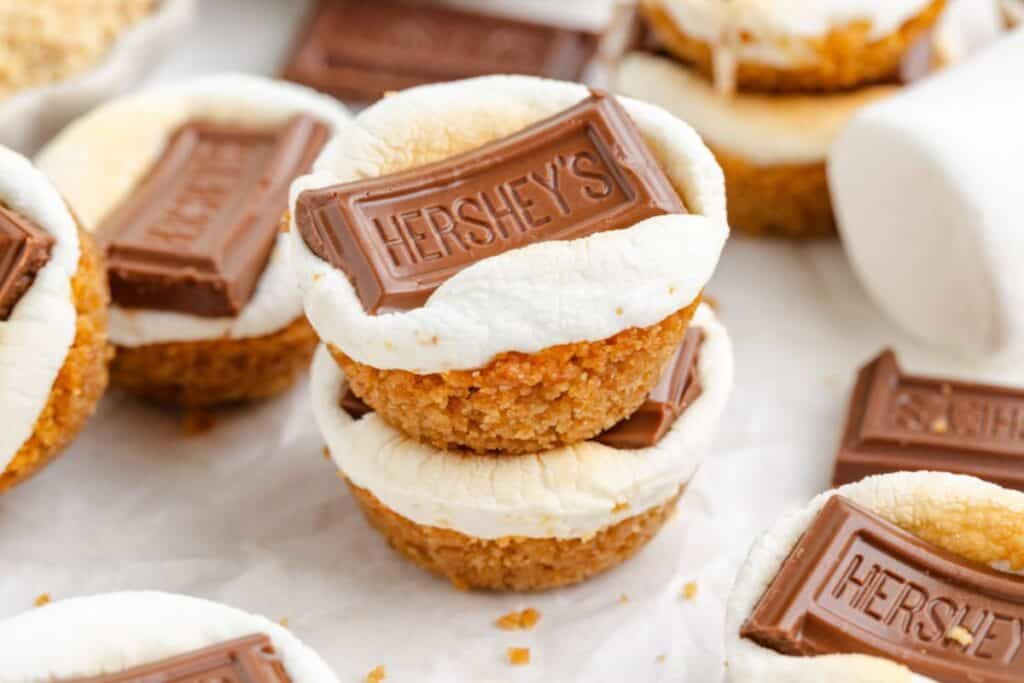 Smores cups topped with toasted marshmallow and Hershey’s chocolate on graham crusts, arranged on a white surface.